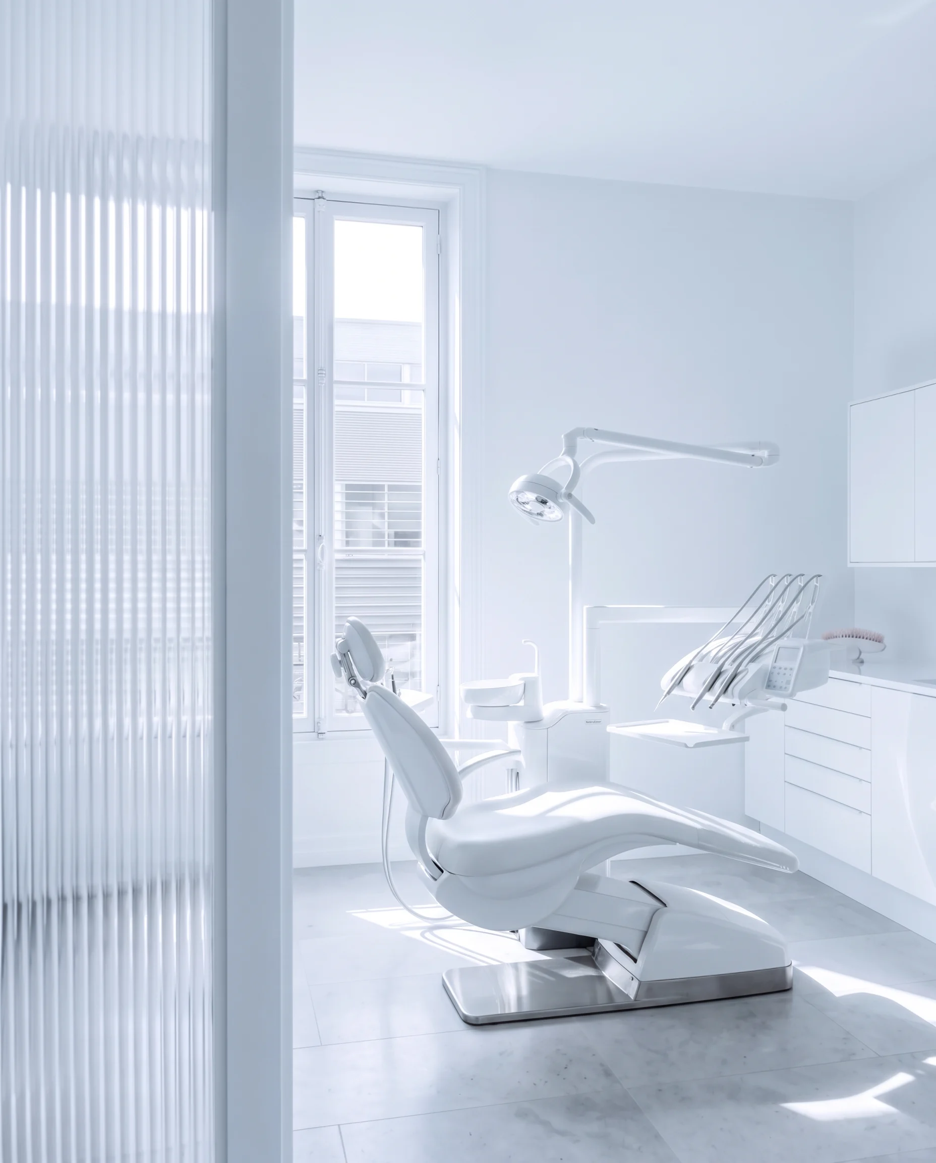 A white treatment room with one chair, a tall Paris window and late-morning sun on the stone floor, seen through reeded glass