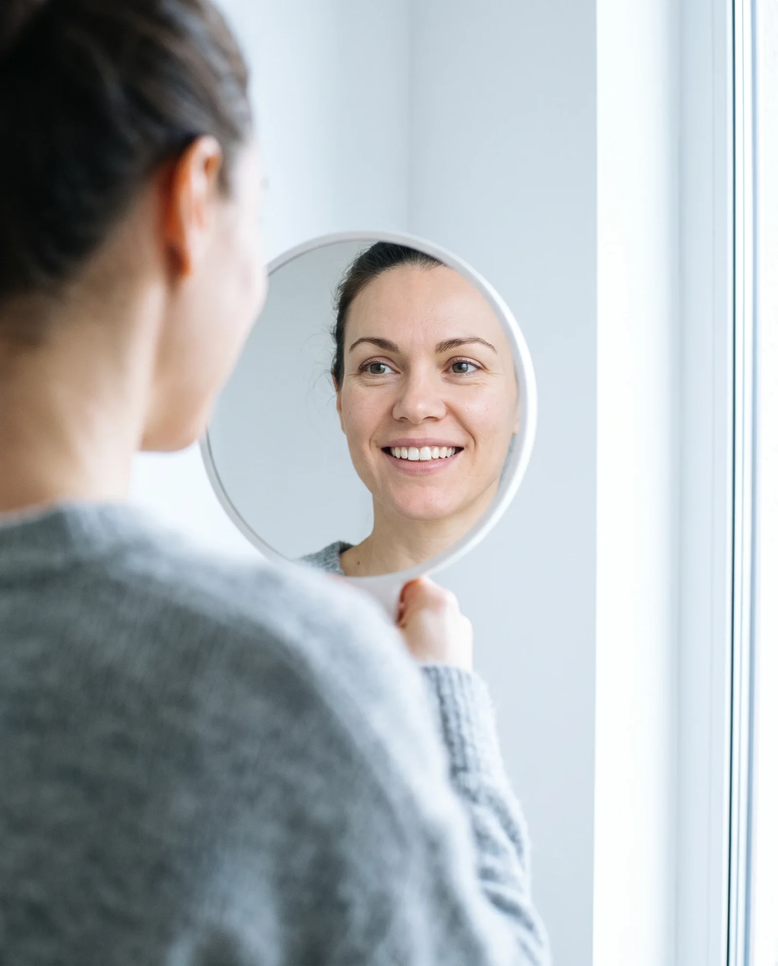 A patient smiling at her reflection in a hand mirror by the window after her fitting